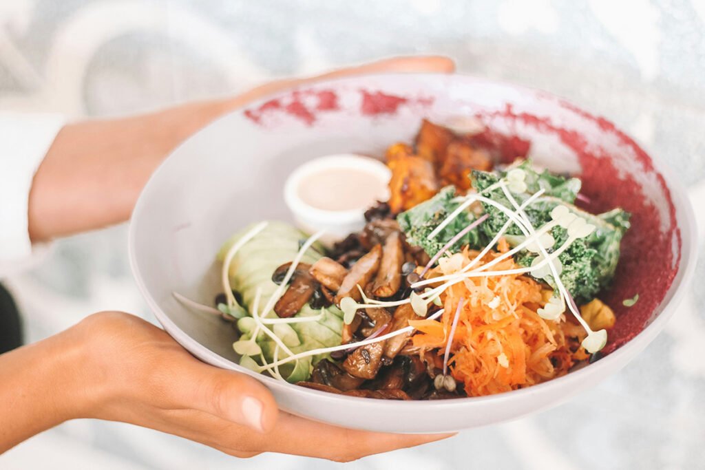 Hands holding a vegan bowl of food with carrots and sprouts.